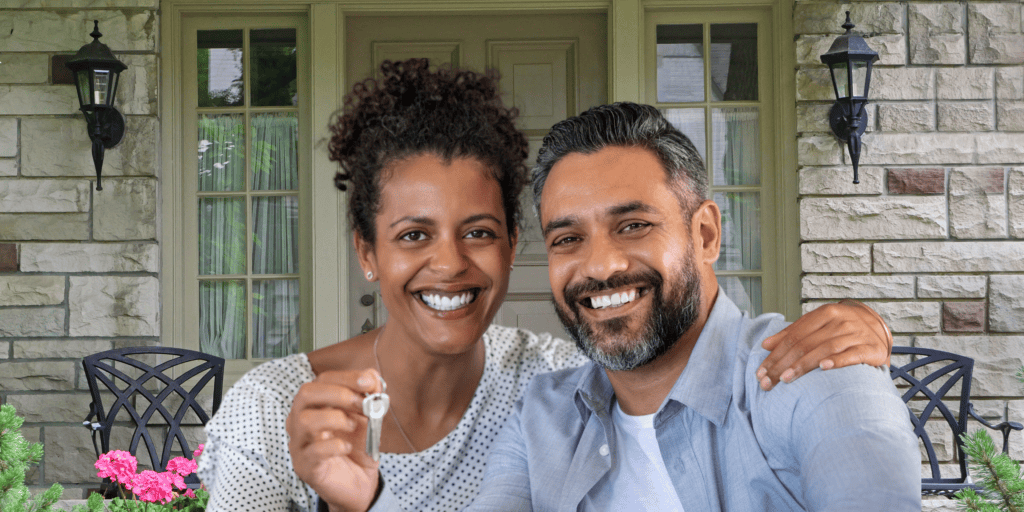 A smiling couple holds a house key outside their stone home, proof you can buy a home after a power of sale.